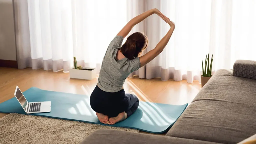 Woman doing yoga next to a laptop in a living room