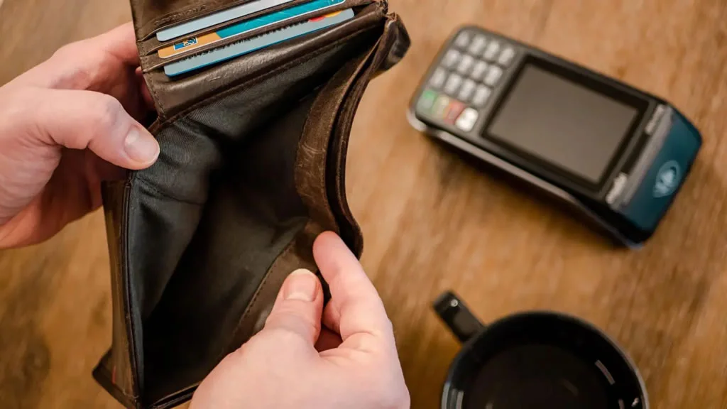 A person holding an opened empty wallet besides a card reader and a coffee mug